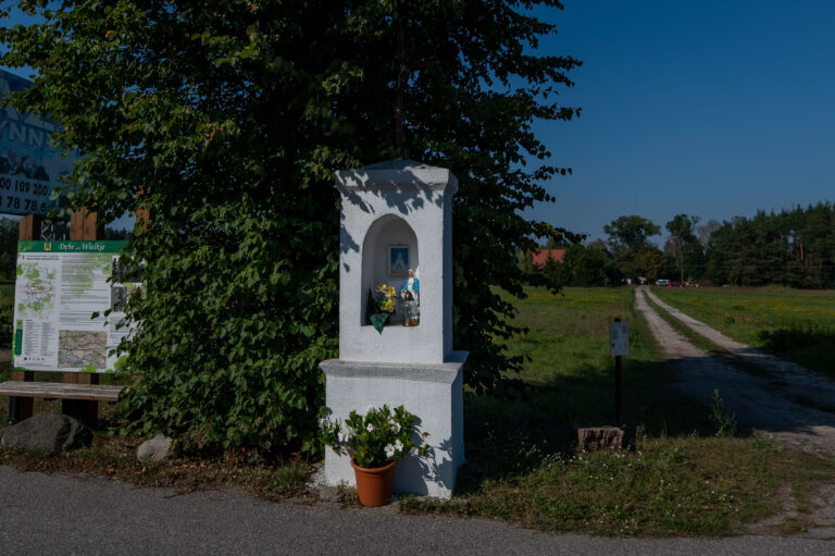 Biała kapliczka zwieńczona metalowym krzyżem z datą 4 września 1907 r. – Świadek bitwy z bolszewikami w 1920 r.